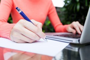 A woman's hands writing notes and using a laptop.