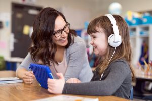 女教师微笑着看着戴着耳机使用平板电脑的年轻女学生。
