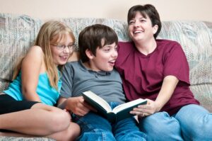 A happy family sits on a couch close together with the mother reading everyone a story out of a large hard bound book.