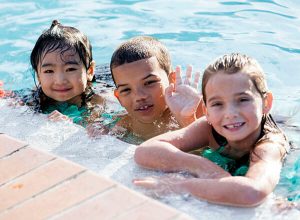 Two girls and a boy in a pool holding on to the wall, the boy in the middle is waving hello.