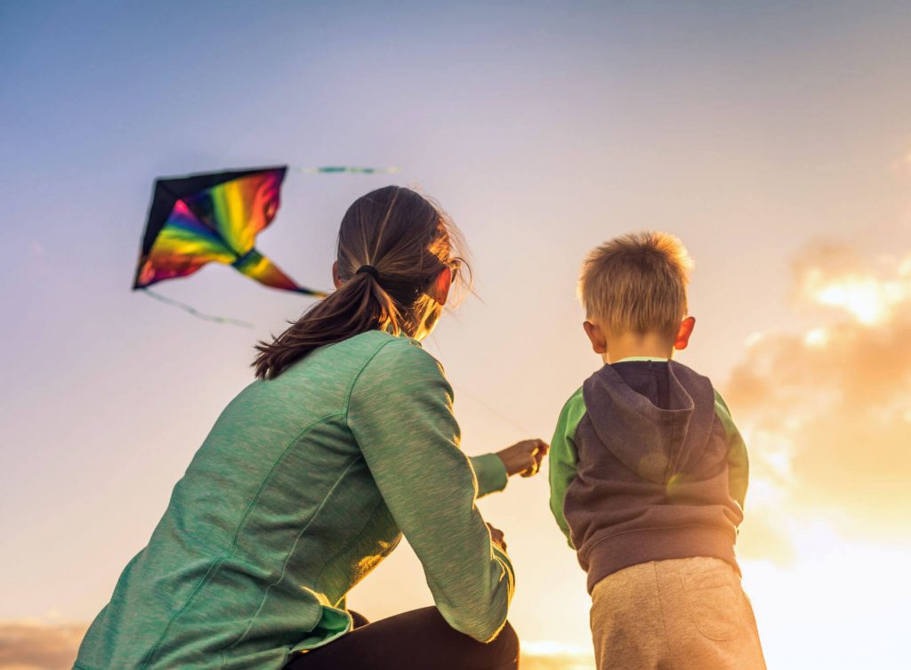 A mother and young son flying a rainbow coloured kite at sunset.