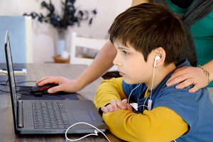 A mother stands behind her son who is sitting a laptop wearing earphones for a telehealth session.