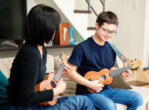 患有唐氏综合症的十几岁男孩与一名辅助工作者坐在家中的沙发上，两人都在弹奏尤克里里琴。