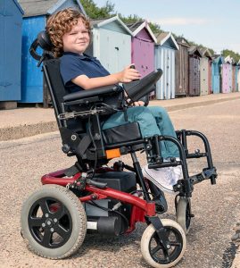 Young boy in power wheelchair by beach huts at the beach.