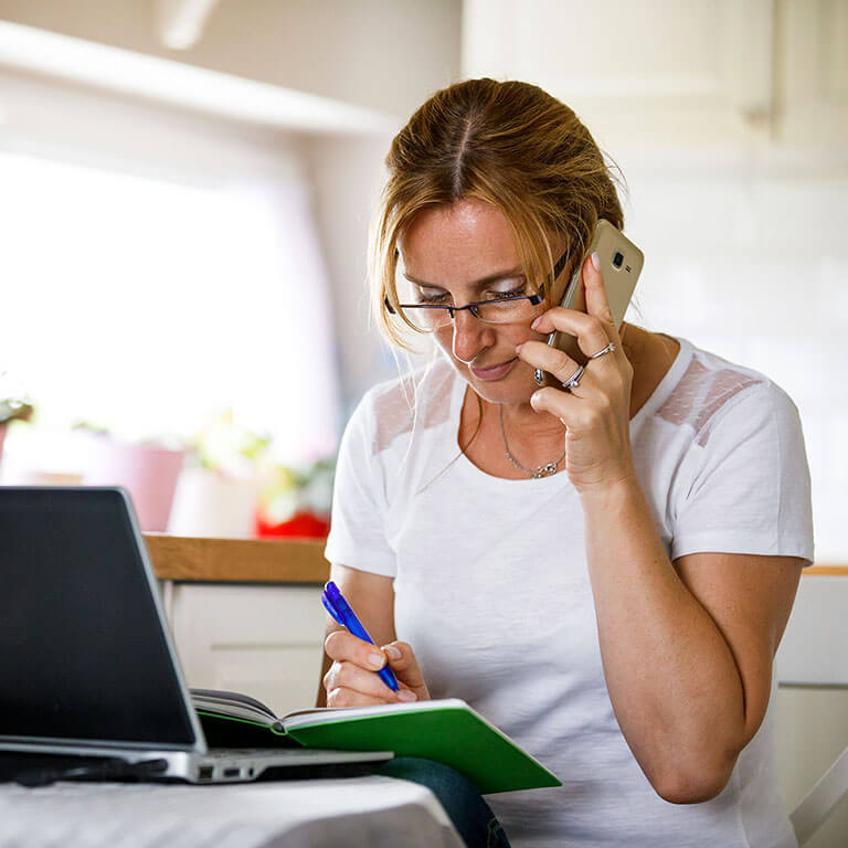 Woman using a laptop while on the phone and writing down notes.