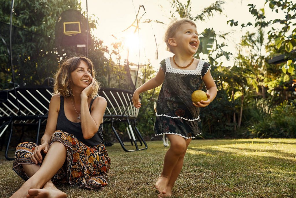 A mum sitting on the grass in the backyard as her young daughter runs around.