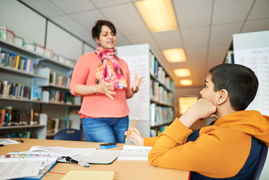 A male student sitting in the school library doing schoolwork, a teacher is standing in front him and they are having a conversation.