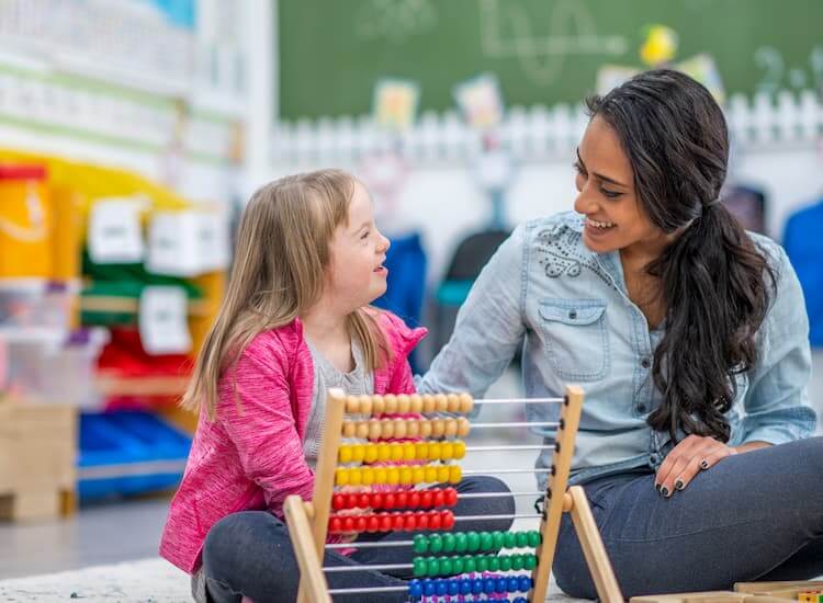 Educator sitting on the floor talking to a young girl with Down syndrome who is playing with an abacus.