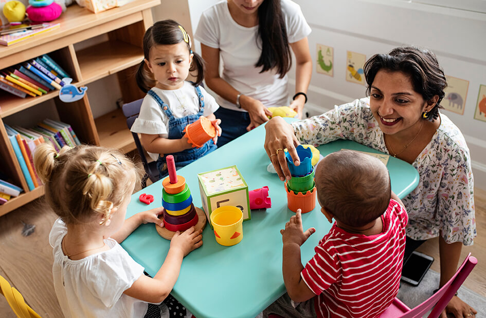 幼儿教育工作者坐在地板上与一名儿童玩叠叠杯，这名儿童与另外两名儿童坐在一张桌子旁。