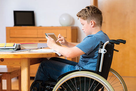 坐在轮椅上的少年坐在厨房的桌子旁，使用电脑平板电脑做功课。