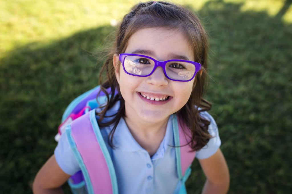 A smiling young girl with purple glasses wearing school uniform and a backpack.