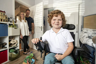 Boy with muscular dystrophy in his bedroom using his powered wheelchair as his mum and dad smiling and watching in the background.