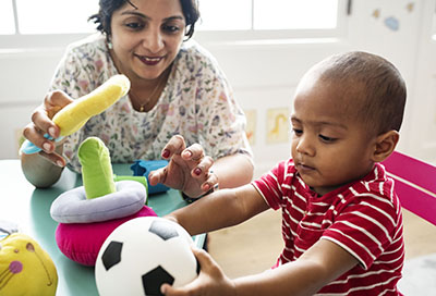 一名小男孩在与幼儿教育工作者玩耍时，伸手拿着一个足球软玩具。