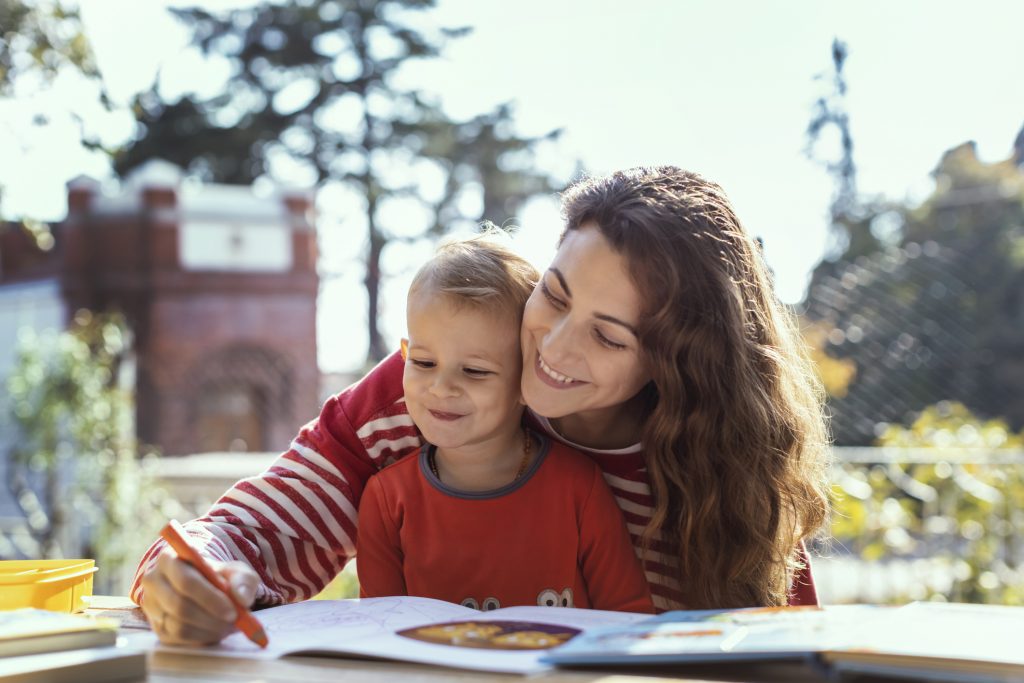 A young mother sits at a park table with a colouring-in book. Her son sits in her lap.