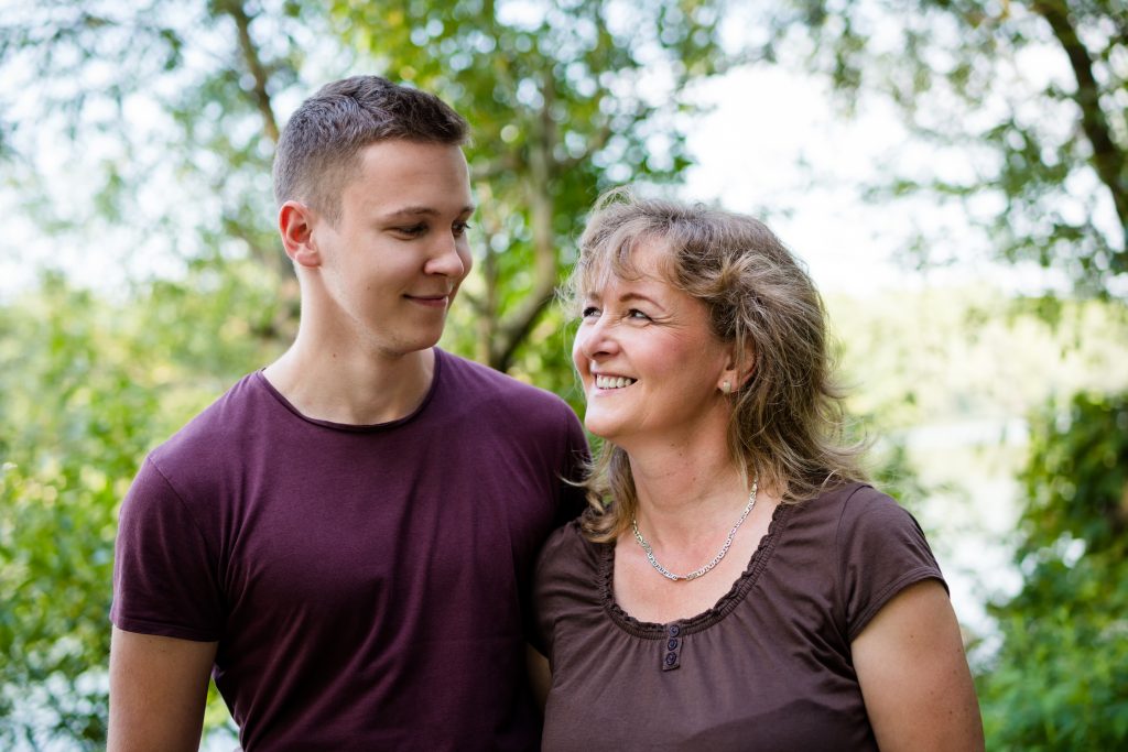 A mother and son stand under a tree. They each have an arm around eachother and are smiling at one another.