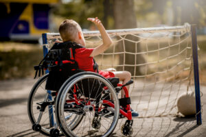 A boy with a disability sits in wheelchair. He plays with a ball next to a football net.