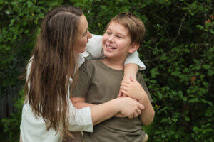 A mother and son embrace and smile to each other, they are standing outside in front of a hedge.