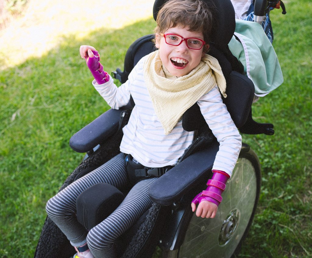 A young girl in a wheelchair wearing glasses, a bib, and wrist guards. She is laughing.