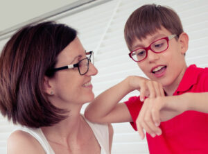 A mother and her son are standing in their lounge room. The boy leans against the blinds and plays with his hands. His mother smiles as she holds and supports him.