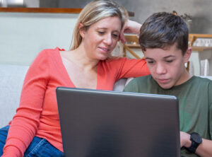 妈妈和儿子坐在沙发上，看着笔记本电脑。妈妈正在辅导他做作业。