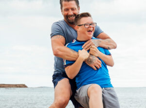 A middle-aged father with a beard, stands behind his teenage son with Down syndrome. He playfully wrestles him on the beach, and they are both smiling.