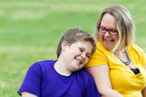 阳光下，一个十几岁的男孩和妈妈坐在草地上。他把头靠在妈妈的肩膀上，两人都在微笑。
