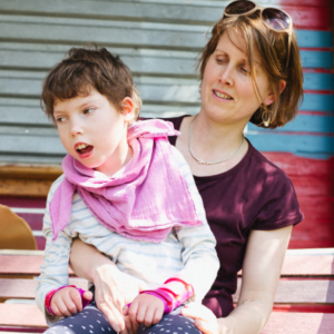 A young school-aged girl sits on her mother's lap outdoors. The girl is wearing a pink big and wrist guards.