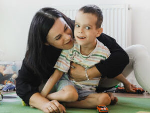 Young boy with cerebral palsy sits on the floor with his Mum. She is hugging him and they are playing with cars.