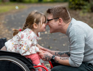 A young girl in a wheelchair is in the park with her Dad. They are playfully holding hands and smiling.