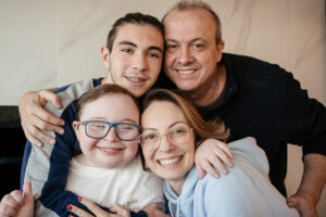 爸爸妈妈和他们的两个儿子在休息室里紧紧拥抱在一起，面带微笑。他们的儿子患有唐氏综合症。