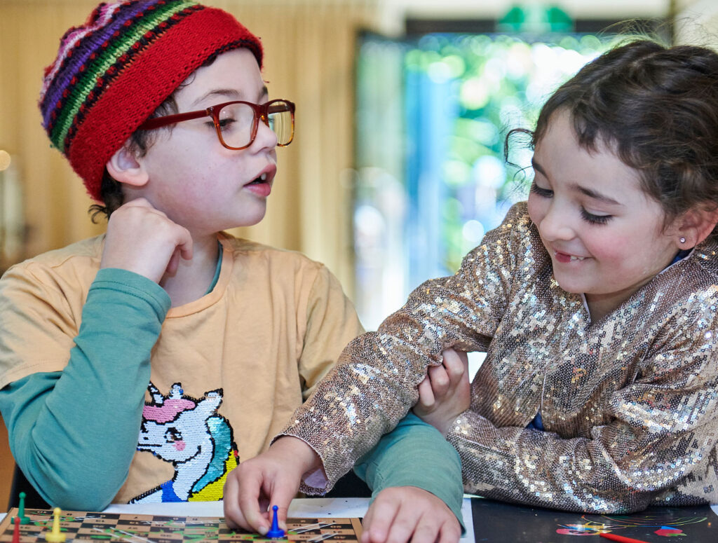 Two children playing a board game together. One child is moving a piece, the other child is looking at them.