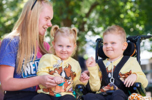 An adult with two young children outsite. One child is sitting on the adult's lap, and the other is in a wheelchair.