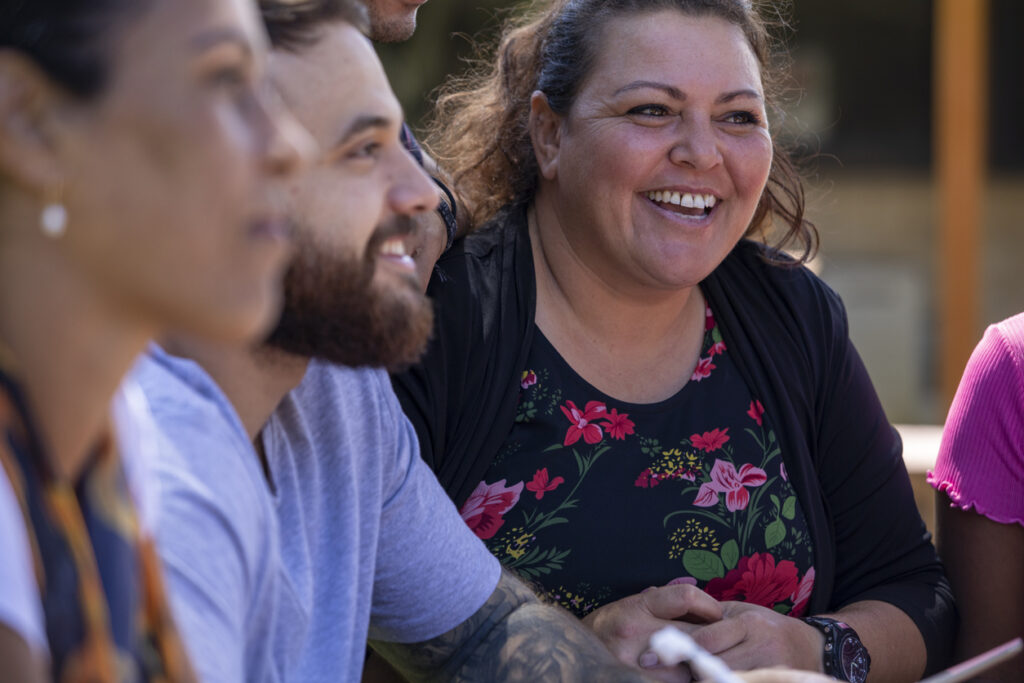 A close-up of a group of Aboriginal adults sitting at an outdoor table. They talk and smile.