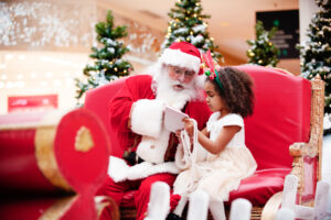 A young girl with curly hair is sitting on a Christmas chair with Santa. They are looking at a gift she was given by Santa.