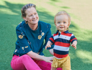 A mother sitting on the grass smiling at a toddler.