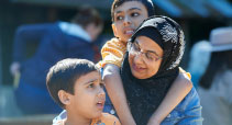 The mother is wearing a hajib and is outdoors with her two autistic boys. One sits next to her and the other is behind with his arms around her shoulders.