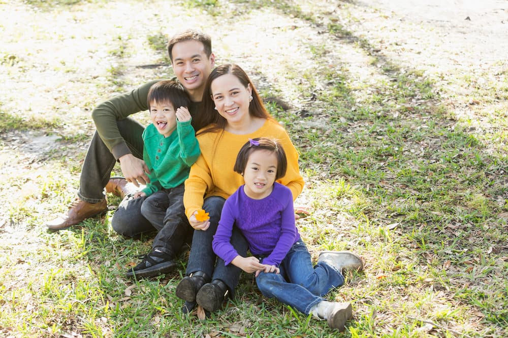 A young Asian family with two school-aged children sits on the grass in the park. They are smiling.