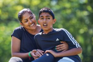 A teenage boy is supported by his mum, and she has her arms around him. They are in the park. He leans toward her, and she smiles lovingly.