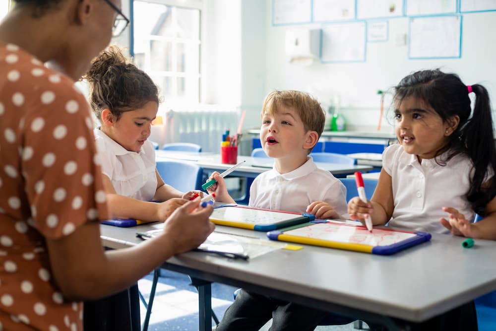 Three primary school students sitting in a classroom using mini whiteboards. The teacher is sitting beside them.