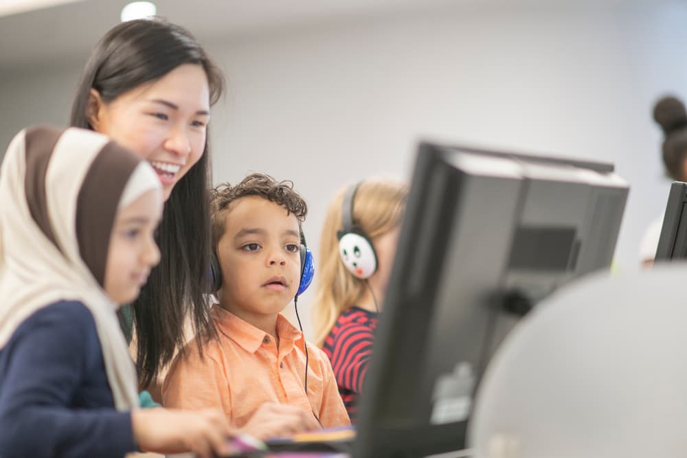 Two young students sit with their teacher looking at a computer monitor.