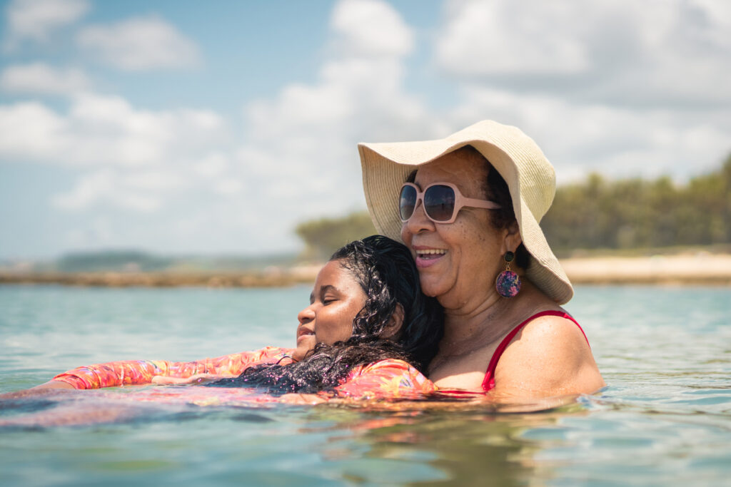 An Aboriginal grandmother is in the sea. She is wearing a sunhat and sunglasses and a red swimsuit. She is holding her granddaughter. They are both smiling.
