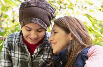 A teenager with a mental health illness sits in the garden with their Mum. The Mum leans on her child and they are both smiling. The teenager wears a beanie and looks down.