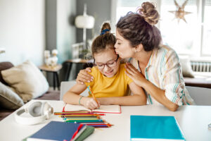 Young girl is sitting at a desk and doing her homework. Her mum sits next to her and gives her a hug and kiss on the cheek.