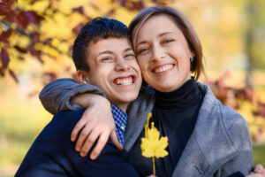 portrait of mother and son in autumn city park, teen boy and woman posing with yellow leaves, they hug, laugh and have fun, bright sunny day
