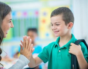 A school boy gives his mother a high-five as she leaves the classroom. Both are smiling.