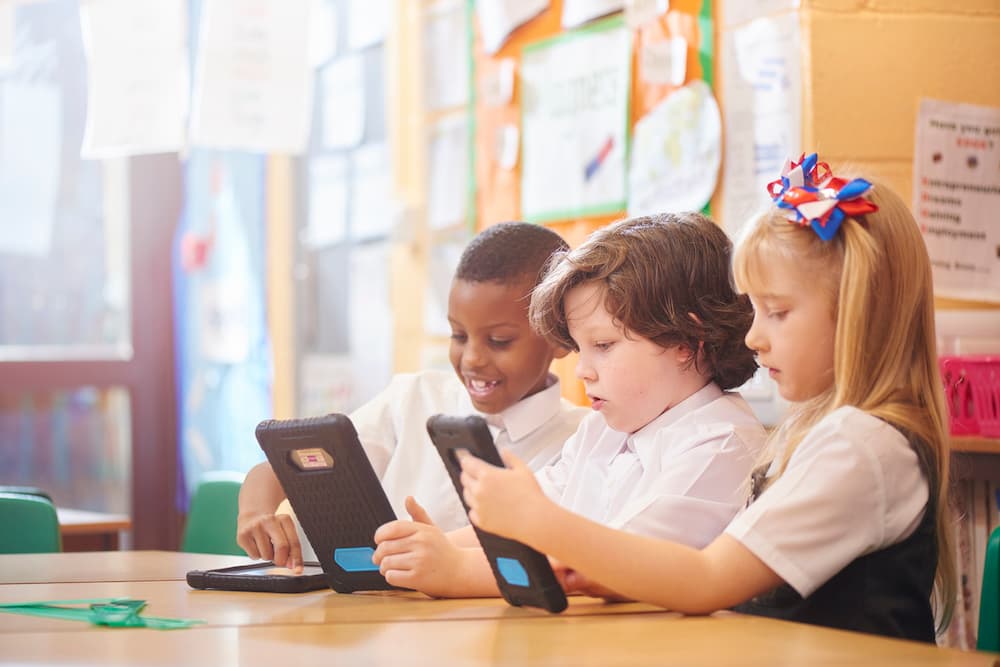 Three primary aged school children sit at their desks working on digital tablets.