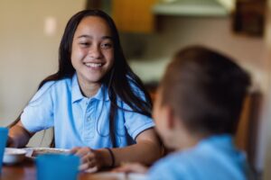 Teen girl having breakfast with her younger brother. They are both at the kitchen table laughing.