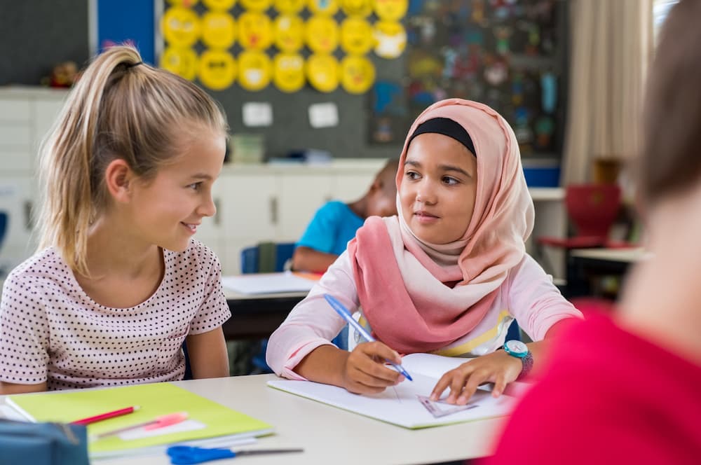 Two school girls sit at a desk working together. They are smiling. One child wears a hijab.