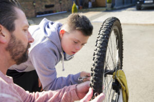 A boy with down syndrome fixes a bike with his father on the driveway.