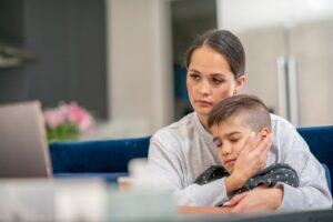 A mother holds her young son on her lap. He looks unwell. She is looking at a laptop.